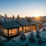Vue aérienne d'une maison de montagne contemporaine entourée de neige et d'arbres, baignée par la lumière chaude du soleil levant.