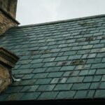 Gros plan sur un toit en ardoise ancien et une cheminée en pierre sous un ciel gris, avec des ardoises patinées.