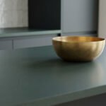 Gros plan sur un plan de travail de cuisine vert olive. Un bol en laiton brossé est posé dessus, avec des meubles sombres et un carrelage blanc en arrière-plan.